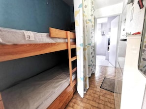 A bunk bed with a wooden frame, a grey mattress, and a plastic cover. There is a curtain with a leaf pattern partially drawn. The room has a tiled floor and a door leading to another room. - Ground floor apartment, close to beach - 4 guests (Cap d'Agde)