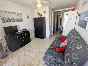 Ein kompakter Wohnraum mit Küchenzeile, einer Couch mit gemusterten Kissen, einem Deckenventilator und einem Segelbootbild.