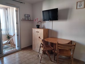 Ein Zimmer mit einem Holztisch, Stühlen, einem an der Wand befestigten Fernseher, einem Schrank und einer Glastür zum Balkon.