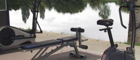 Outdoor gym equipment including a bench, dumbbells, and an elliptical machine, with a calm lake and greenery in the background.
