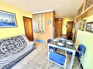 A dining area with a table set for four, a sofa with a patterned cover, and a kitchen in the background. - Comfortable 2-bedroom apartment 100m from the beach, air conditioning, balcony, satellite TV. (Roses)