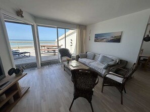 Ein Wohnzimmer mit einer Couch, Sesseln, einem Couchtisch und einem Fernseher. Es gibt einen Balkon mit Blick auf den Strand.
