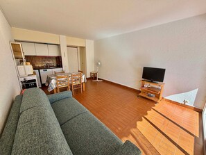 Ein Wohnzimmer mit Sofa, Fernseher auf einem Holzschrank, Essbereich mit Tisch und Stühlen sowie Küchenzeile mit Geräten.