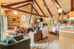 A wooden cabin interior with a dining area, sofa, kitchen, and a large window.