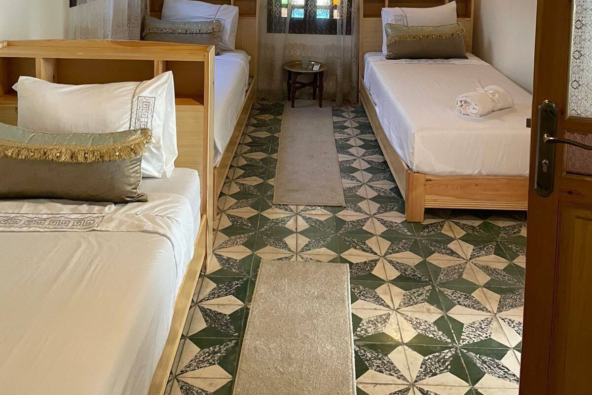 A room with two beds, a small table, a decorative window, and a patterned floor.