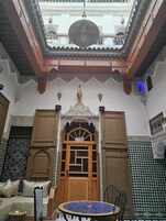 Interior tradicional marroquino com porta de madeira, mesa estampada e vista do pátio.