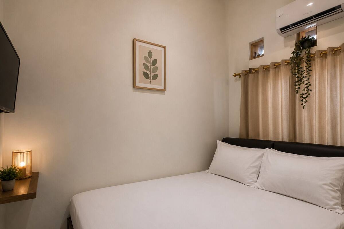 A neatly made bed with white linens, a framed leaf artwork on the wall, a bedside table with a lamp, and a hanging plant.