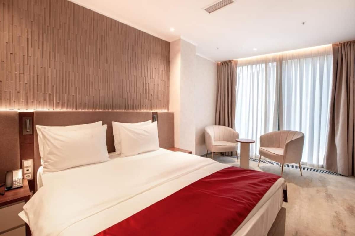 A hotel room with a large bed, a bedside telephone, a small table, two chairs, and a window with curtains.