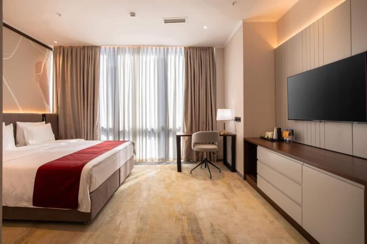 A hotel room with a bed, a desk, a chair, a TV, and a large window with curtains.