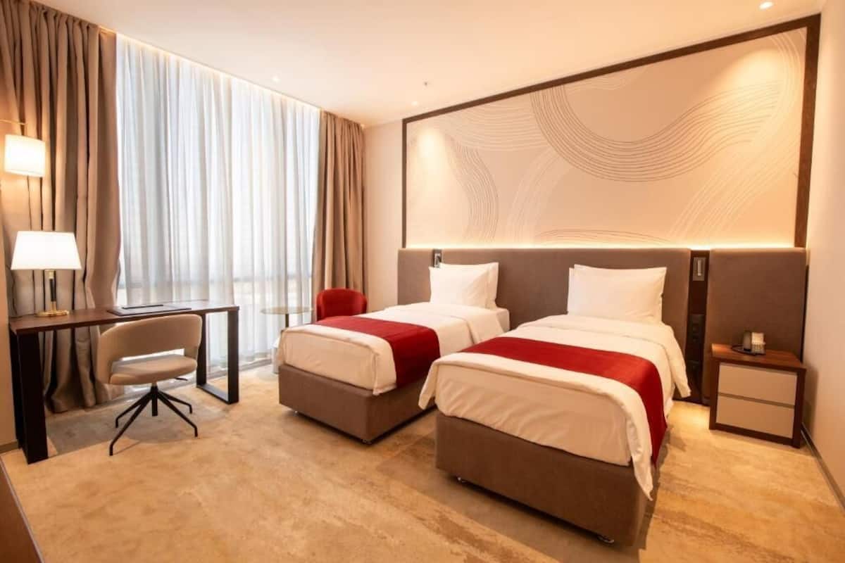 A hotel room with two beds, a desk, a chair, a nightstand, and a large abstract wall mural.