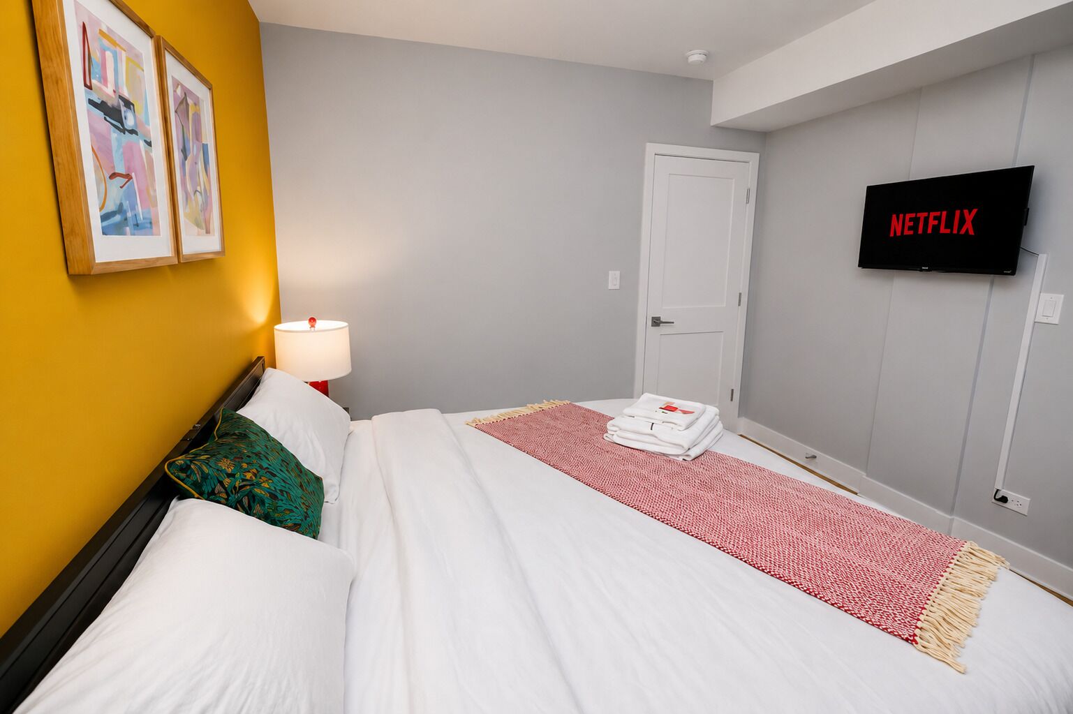 A hotel room with a bed, a TV displaying 'NETFLIX,' a door, and two framed artworks on the wall.