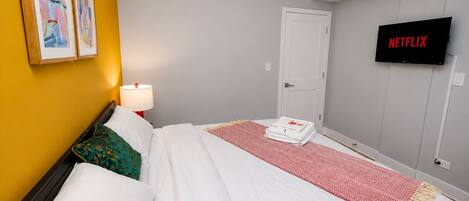 A hotel room with a bed, a TV displaying 'NETFLIX,' a door, and two framed artworks on the wall.
