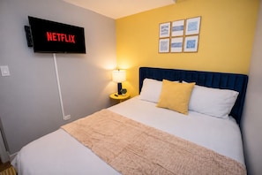 A hotel room with a bed, a TV displaying 'NETFLIX,' a lamp, and framed artwork on the wall.