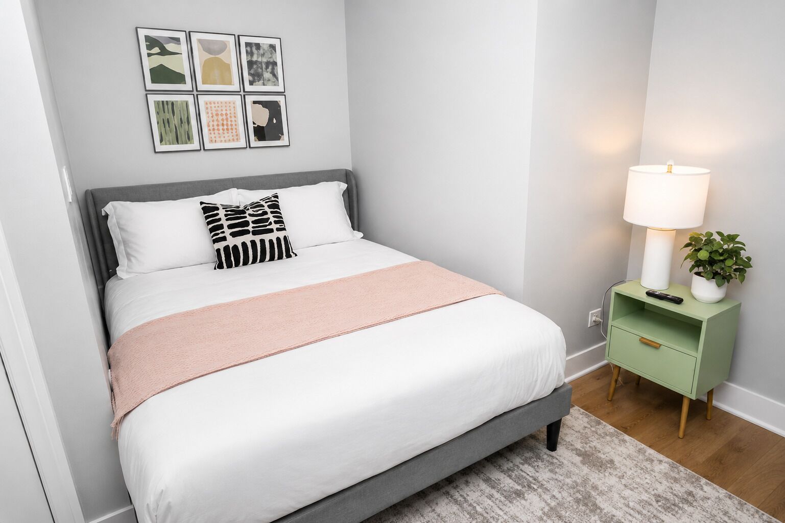 A neatly made bed with a grey headboard, a white and pink bedspread, and a grey pillow. There is a green nightstand with a lamp, a remote, and a potted plant.