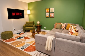 A living room with a grey sofa, a green armchair, a coffee table, a TV mounted on the wall, and a green wall with four framed pictures.