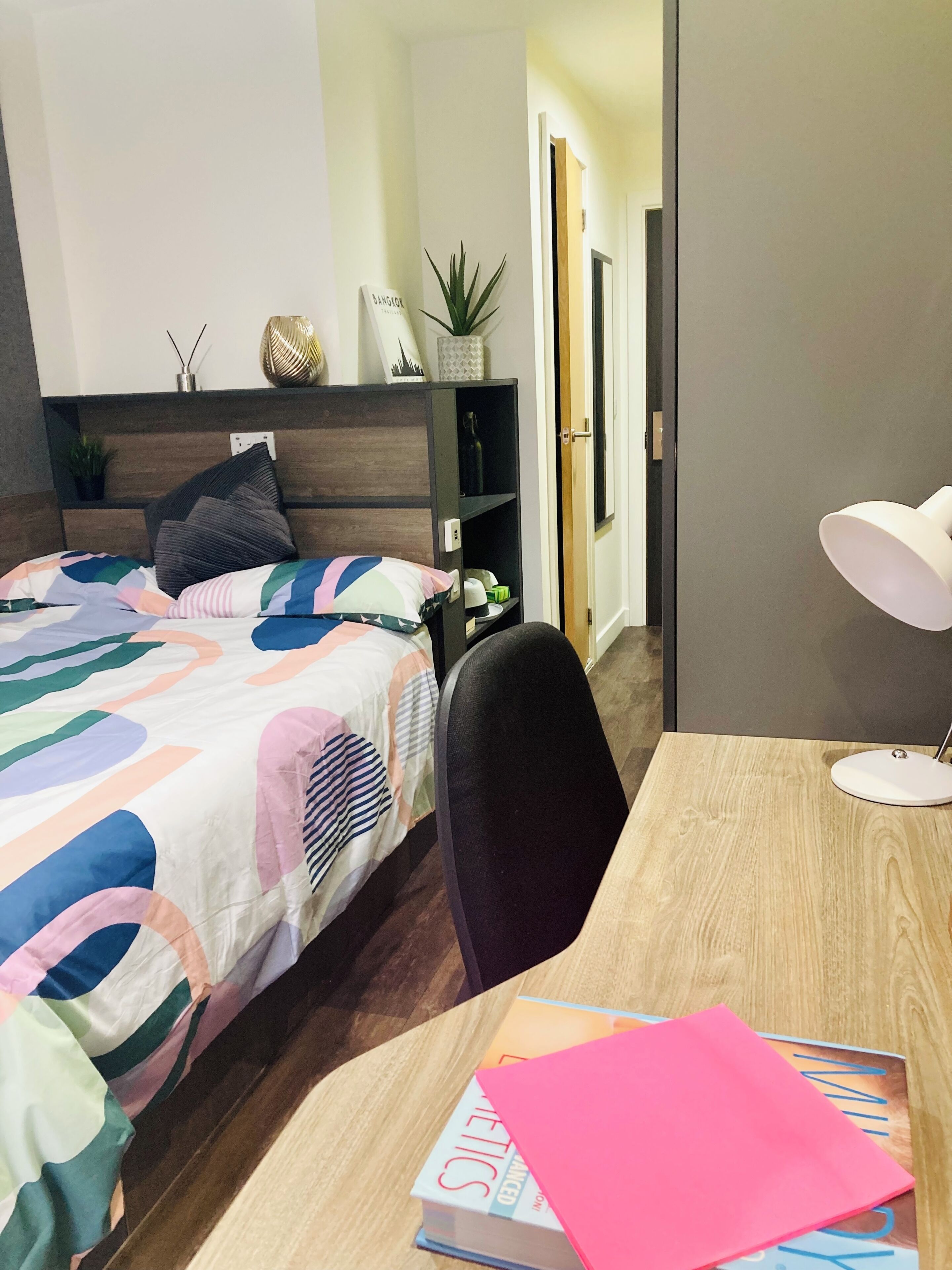 A bedroom with a bed, a desk, a chair, a bookshelf, a potted plant, and a door.