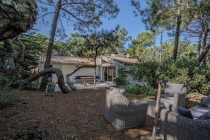 Uma casa com telhado de telhas, um pátio com móveis de vime e um jardim com pinheiros.
