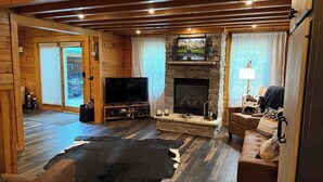 A log cabin interior with a stone fireplace, a television, leather furniture, and a large fur rug.