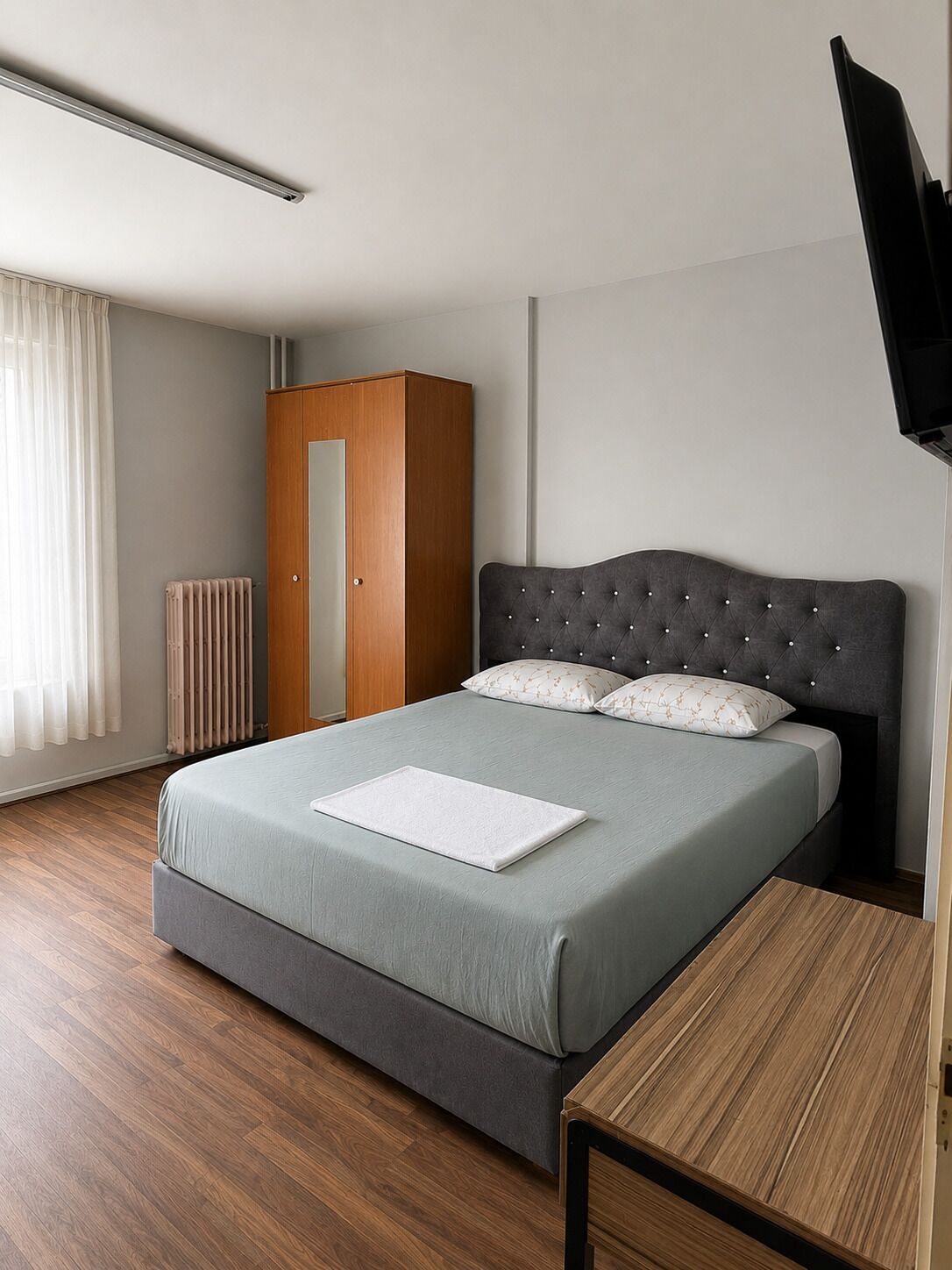 A modern bedroom with a bed, a wooden wardrobe, a radiator, and a TV mounted on the wall.