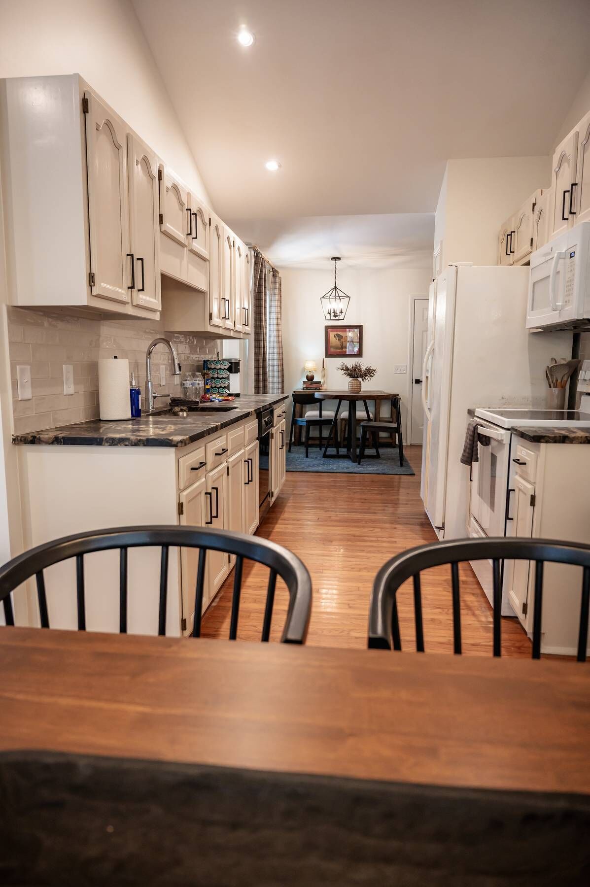 A modern kitchen with white cabinets, a dining area with a table and chairs, and a hanging light fixture.