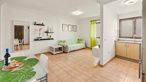 Een woonkamer met een bank, een eettafel met een fles en lekkernijen, een keuken met kastjes en een spoelbak, en een hal met een spiegel en een deur.