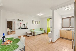 Ein Wohnzimmer mit einer Couch, einem Esstisch mit Flasche und Snacks, einer Küchenzeile mit Schränken und Spüle sowie einem Flur mit Spiegel und Tür.