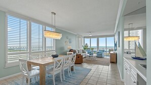 A spacious living area with a dining table, white chairs, a sofa, and a view of the ocean through large windows.
