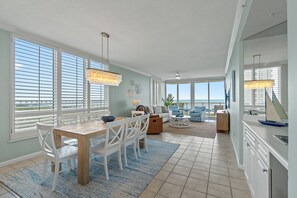 Een ruime woonkamer met een eettafel, witte stoelen, een bank en uitzicht op de oceaan door grote ramen.