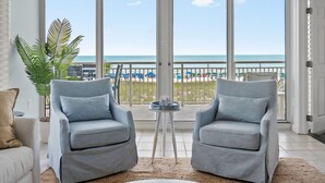 A room with a balcony overlooking the beach, featuring two grey armchairs, a white sofa, and a small table with a plant.