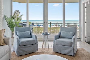 Een kamer met een balkon met uitzicht op het strand, met twee grijze fauteuils, een witte bank en een kleine tafel met een plant.