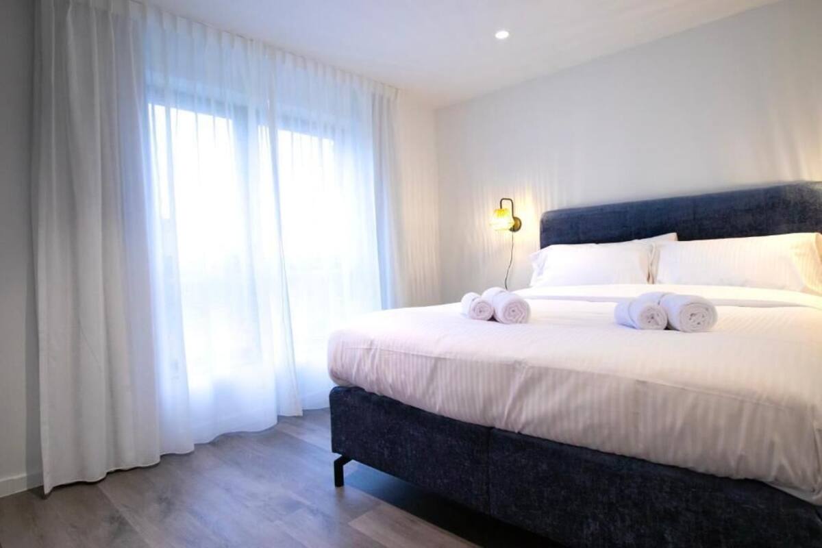 Un lit bien fait, avec des serviettes pliées, une tête de lit sombre et un luminaire fixé au mur.