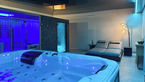 A modern spa room with a hot tub, a massage table, and a glass-enclosed sauna.
