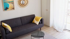 Ein modernes Wohnzimmer mit einem grauen Sofa, einem runden Spiegel, einer Korbtasche als Lampe und einem gerahmten Bild an der Wand.