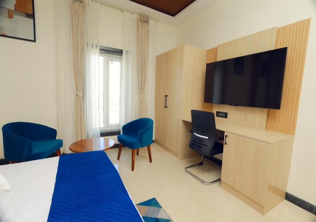 A hotel room with a bed, two chairs, a desk, a TV, and a window with curtains.