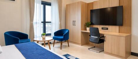 A hotel room with a bed, two blue chairs, a desk with a TV, and a window with curtains.