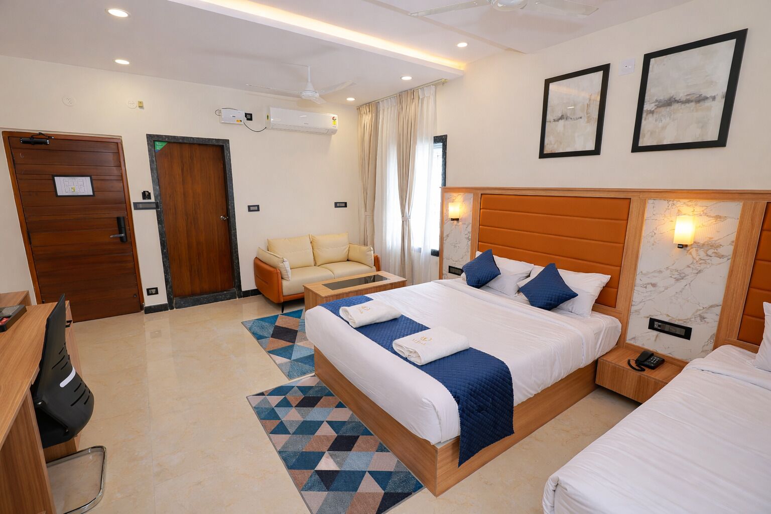 Une chambre d’hôtel avec un grand lit, un canapé, un bureau et une chaise. Il y a deux unités de climatisation au mur, un ventilateur de plafond et une fenêtre avec des rideaux.