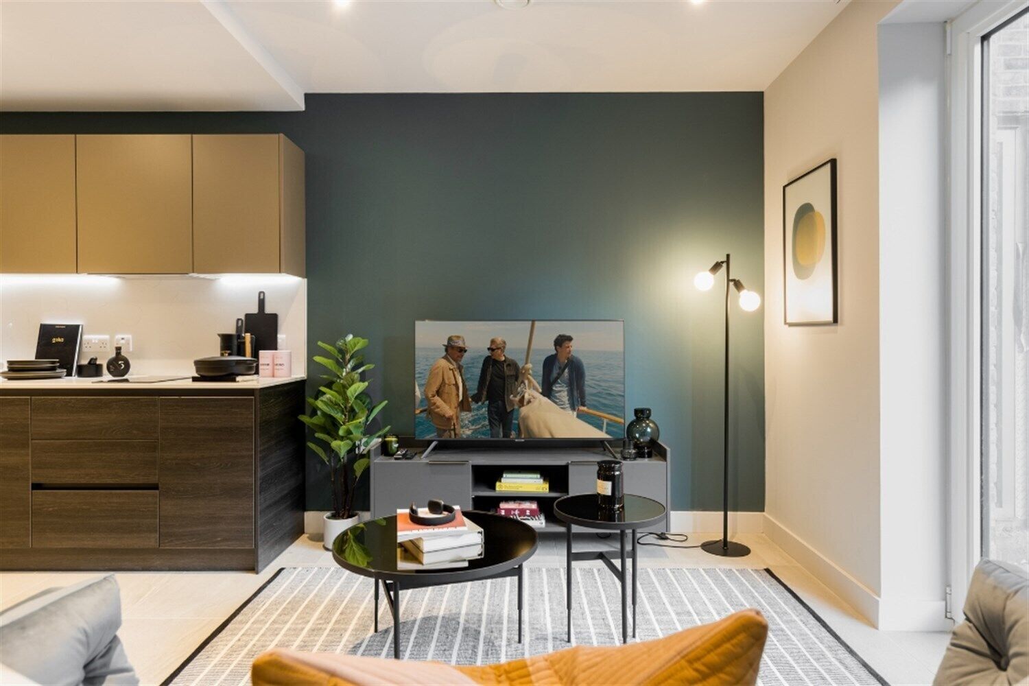 A modern living room with a flat-screen TV, a coffee table, a sofa, a kitchen area, and a large window.
