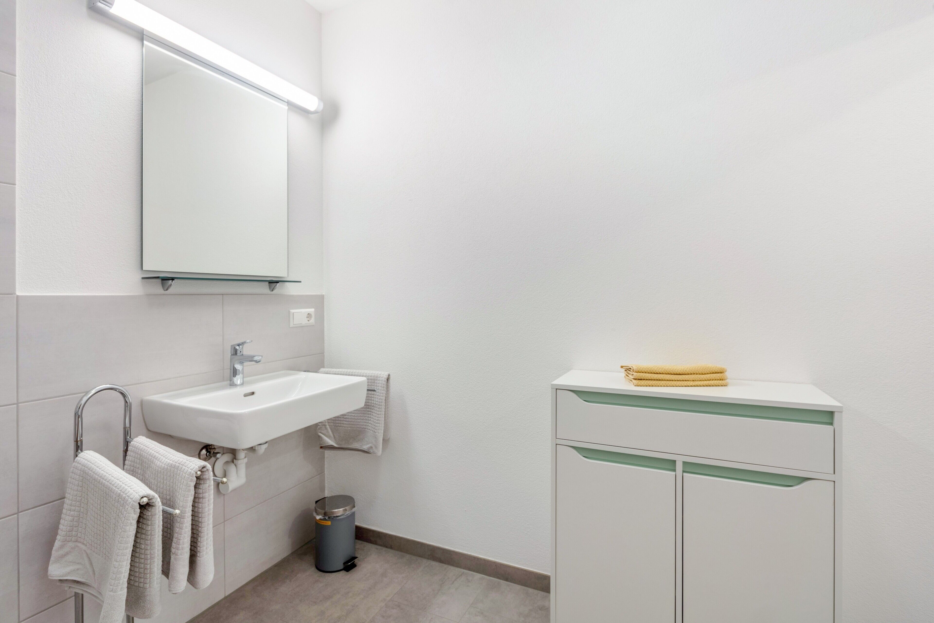Un baño con lavamanos, espejo y mueble blanco, además de toallas y un cesto de basura.