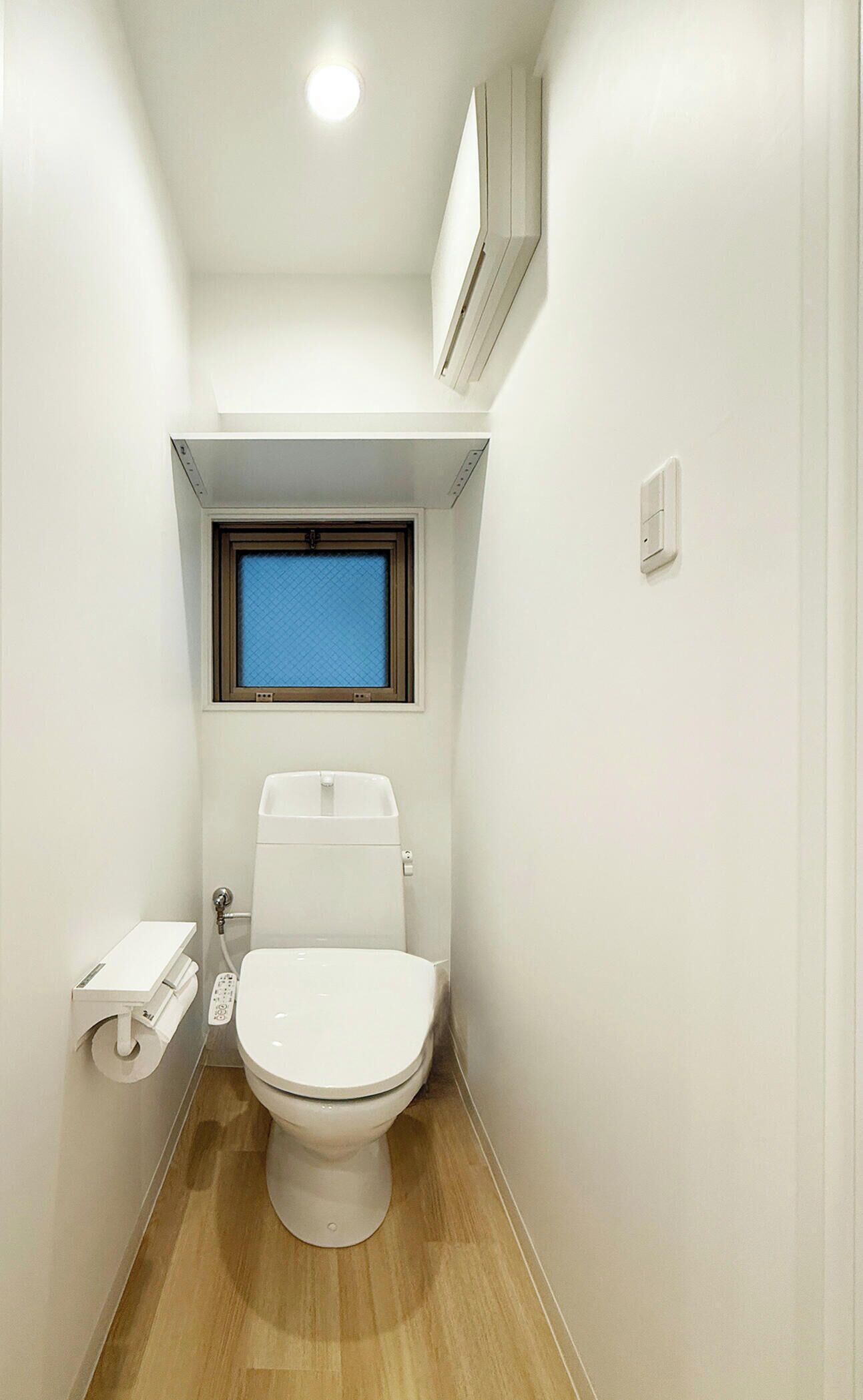 A small, narrow bathroom with a toilet, a window, and a wall-mounted air conditioner.