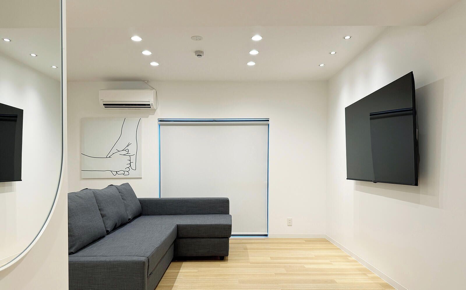 Une pièce moderne avec un canapé d’angle gris, un téléviseur à écran plat fixé au mur, un éclairage encastré au plafond et un appareil de climatisation.