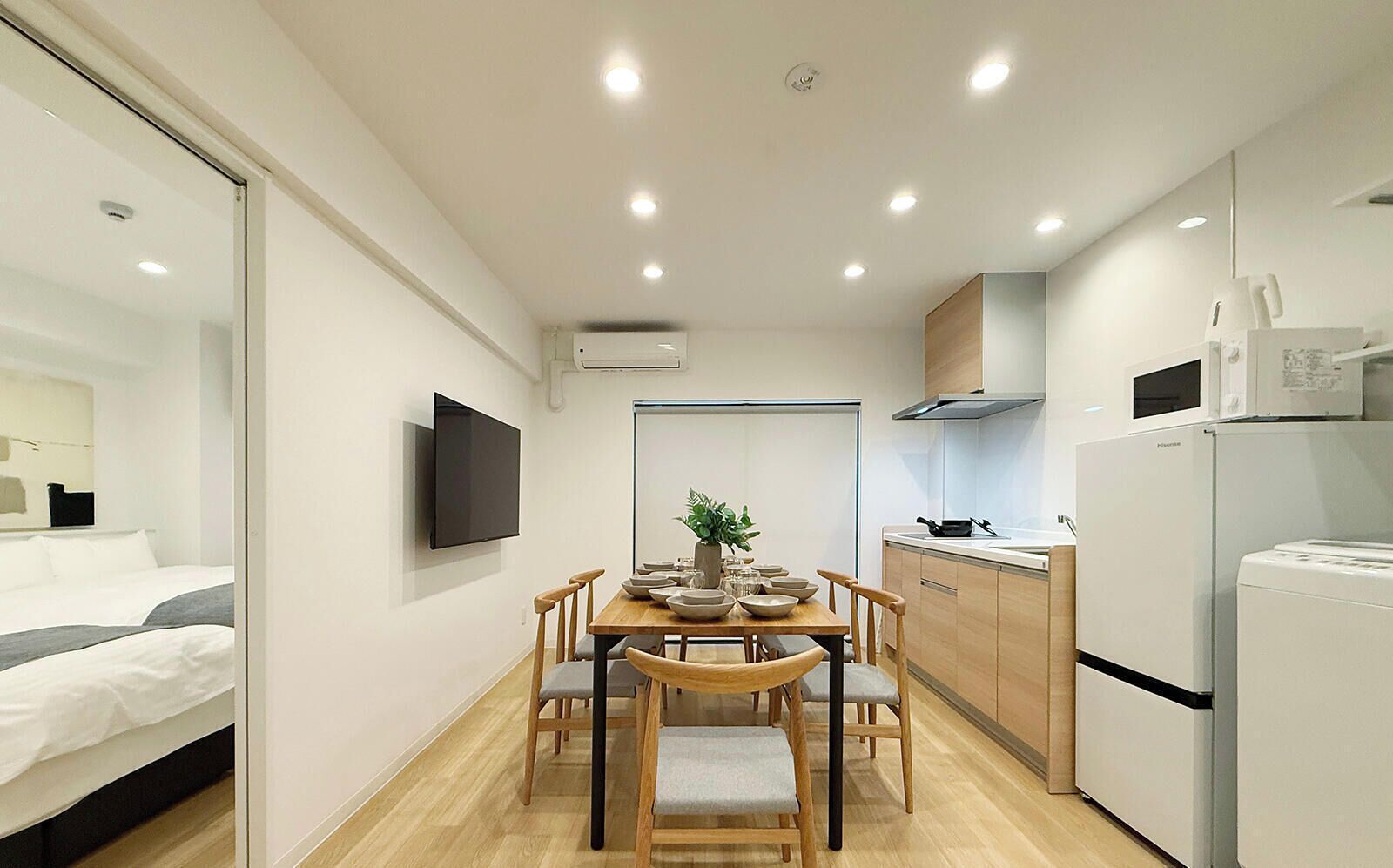A compact hotel room with a bed, a TV, a dining table with chairs, a kitchen area with a fridge, and a microwave.