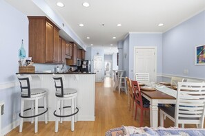 A modern kitchen with wooden cabinets, a central island with bar stools, and a dining area with a wooden table and chairs. - Big Blue - Second Row Home with Ocean Views! (Carolina Beach)