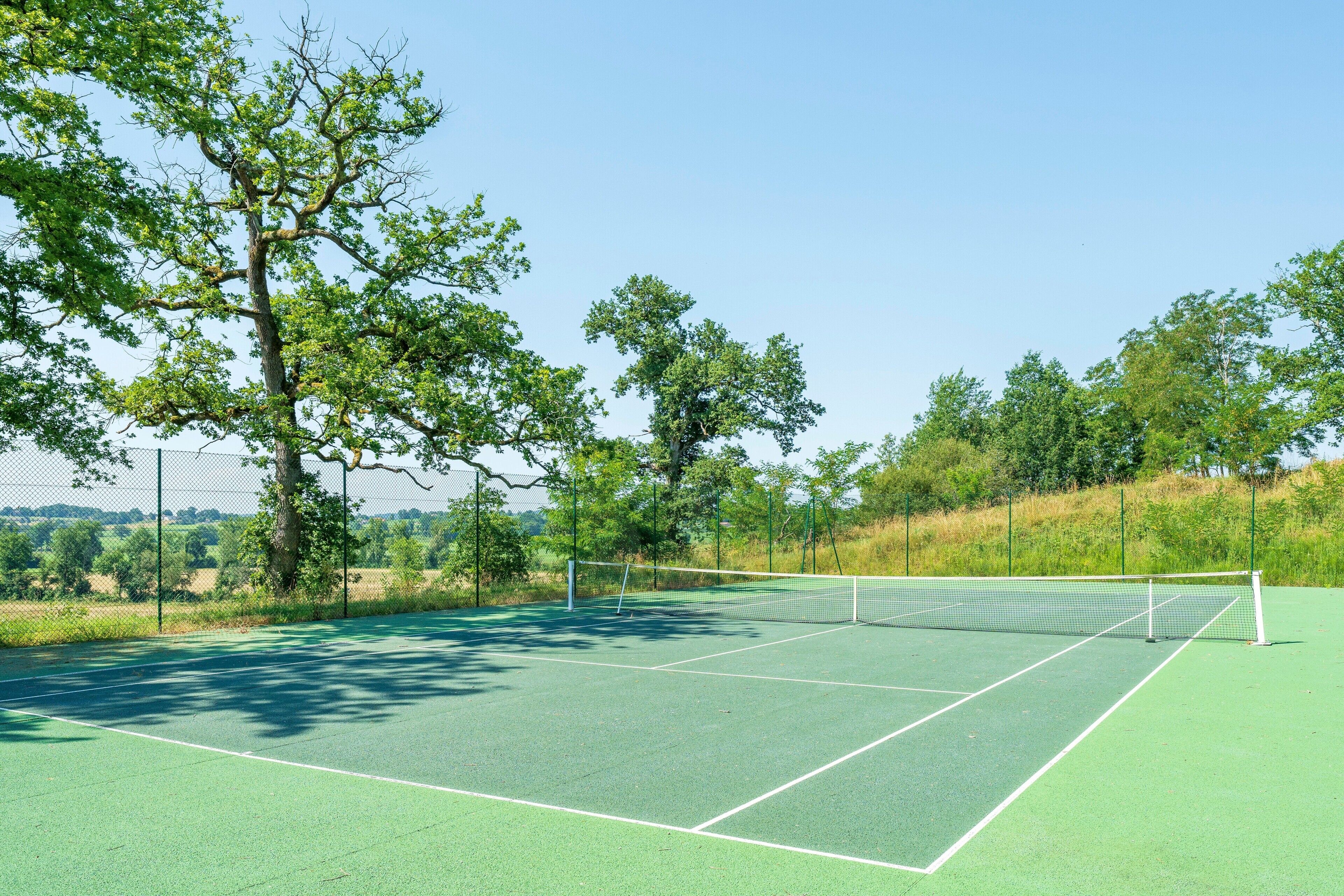 Cancha de tenis al aire libre con red, rodeada de árboles y una valla.