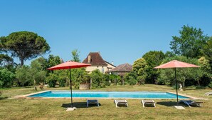 Una piscina con sillas de descanso y sombrillas, rodeada de follaje y una casa al fondo.