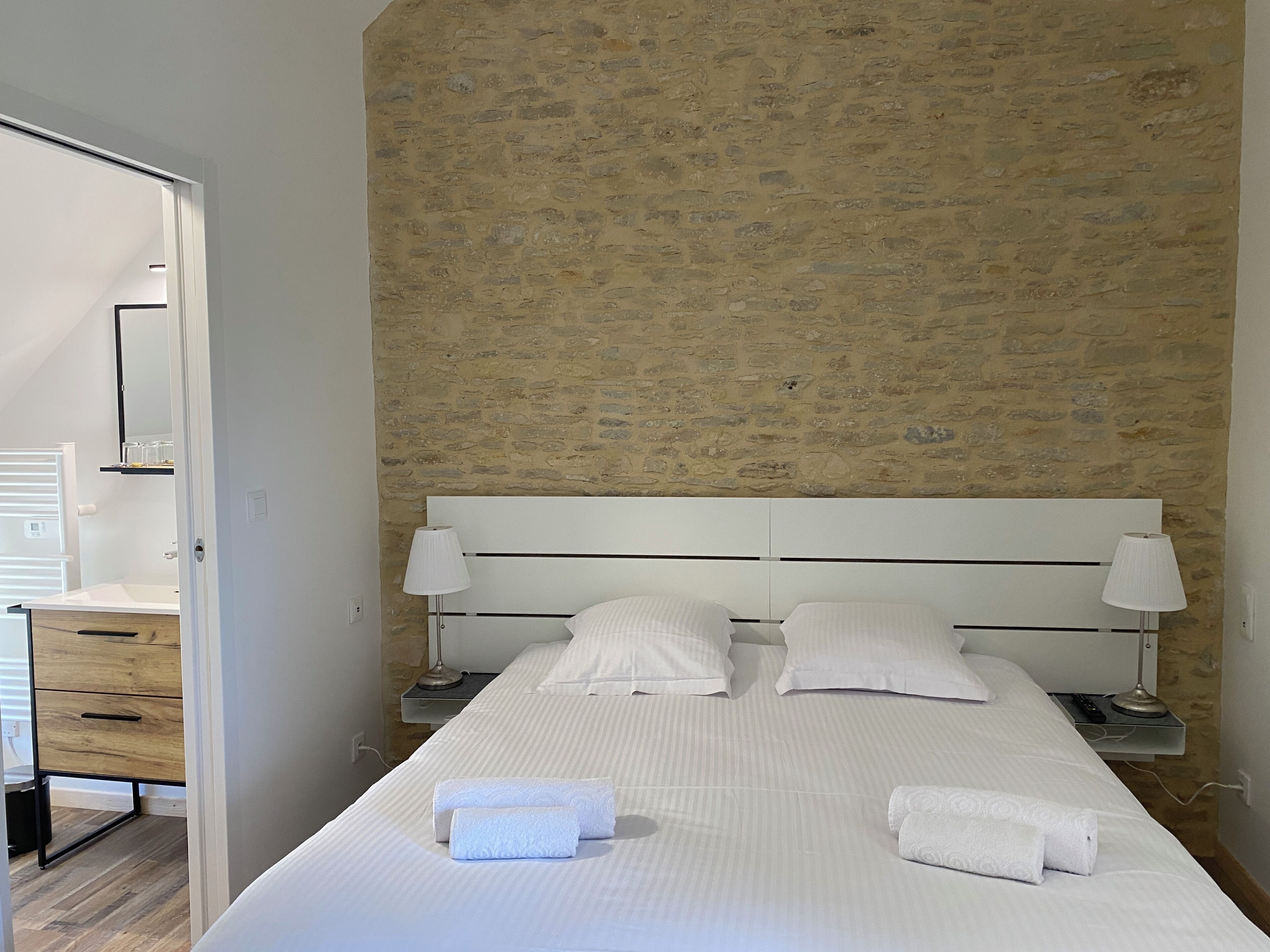 Un dormitorio con una cama grande, dos mesitas de noche con lámparas, un aparador de madera y una pared de piedra.