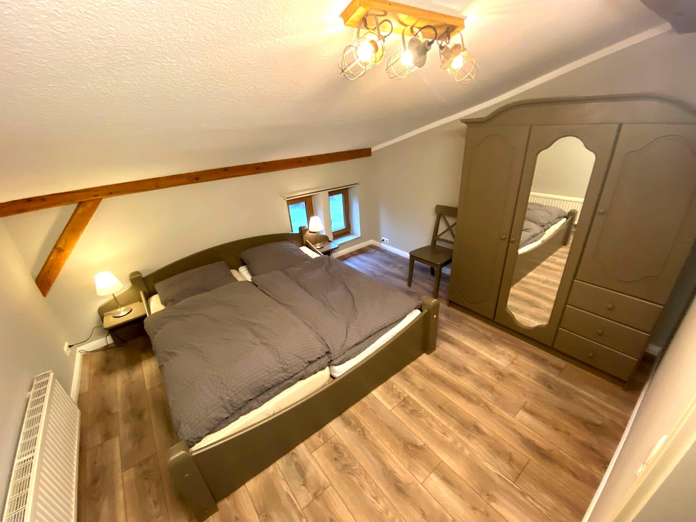 Un dormitorio con cama, mesita de noche, lámpara, ventana, silla, armario y espejo.
