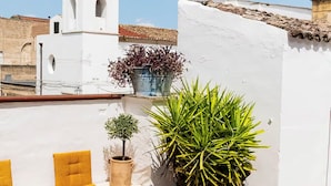 Una terraza en la azotea con un muro blanco, una torre campanario y plantas en macetas.