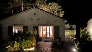 Uma casa com um caminho de pedra que leva à entrada, uma pequena varanda com plantas e o interior iluminado visível pelas portas de vidro.