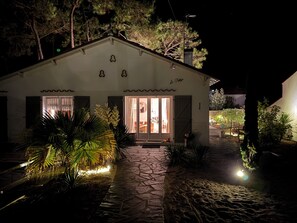 A house with a stone pathway leading to the entrance, a small porch area with plants, and a lit interior visible through the glass doors. - Charming villa 'Le Refuge' heated pool 300m from the beach in a quiet location (La Tranche-sur-Mer)