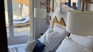 Quarto de hotel com cama, almofadas, mesa de cabeceira, abajur, espelho e janela com cortinas.
